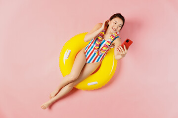 Top view full size young woman of Asian ethnicity in striped swimsuit lies on inflatable ring in pool do selfie shot on mobile cell phone isolated on plain pink background. Summer vacation sea concept