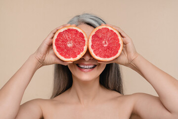 Smiling mature middle-aged woman with bare shoulders naked holding grapefruit covering her eyes isolated in beige background. Beautification and healthy dieting concept