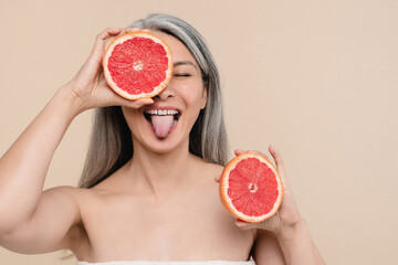 Skin and hair care. Smiling mature middle-aged woman with bare shoulders naked holding grapefruit covering her eyes isolated in beige background. Beautification and healthy dieting concept