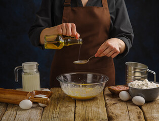 The chef is preparing the dough. Pour olive oil into a glass bowl. Ingredients for dough on the kitchen table. Dark background. Recipes for preparing dishes from dough - pizza, pie, pasta, bread.