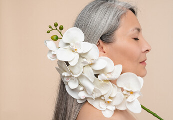 Skin and hair care. Anti-age anti-wrinkle beauty products cosmetics. Beautification and rejuvenation. Closeup photo of mature middle-aged woman holding orchid flower on her face with eyes closed - obrazy, fototapety, plakaty