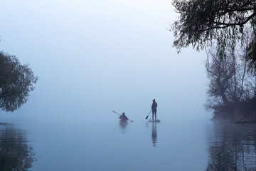 Rear view of two women paddling kayak and stand up paddle board (SUP) at autumn misty river at foggy autumn morning
