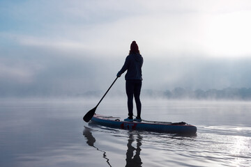 Silhouette of woman rowing stand up paddle board (SUP) at autumn misty and foggy morning at Danube river