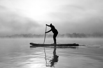 Black and white image of silhouette of woman rowing stand up paddle board (SUP) at misty and foggy morning at Danube river