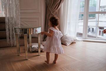 Little baby girl walking barefeet on floor in room. Girl from back in white dress doing her first steps. one child indoors.