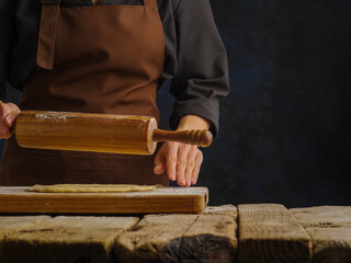 The chef rolls out the dough with a rolling pin on a wooden kitchen table, dark background. Minimalism. The concept is the preparation of products from dough - pizza, pasta, pie. Book of recipes.