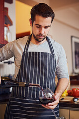 Improve your cooking - add wine. Shot of a happy young man pouring himself a glass of wine while cooking in his kitchen at home.