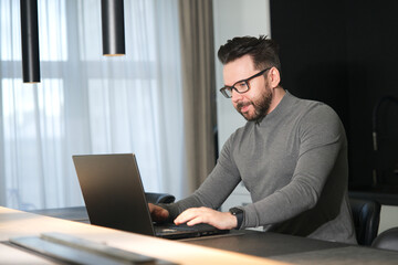 Portrait of serious concentrated focused middle aged bearded man freelancer in glasses is working on his laptop computer, typing on keyboard at home at luxury apartment. Freelance distant job concept.
