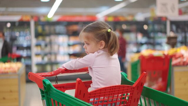 Child, little girl 5 years sitting shopping cart in supermarket while mother choose goods to buy it Female kid shopping cart food store or supermarket. Little kid going shopping. Healthy food for kids