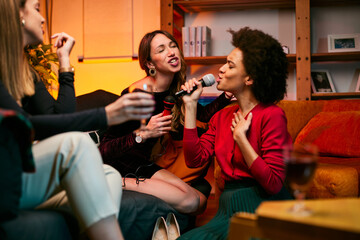 Two multicultural young women having fun at a home party while singing karaoke for their friends.