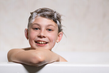 Little, beautiful girl, baby bathes sitting in a white bath with soap suds, shampoo and washes her head with hair. Model photography.