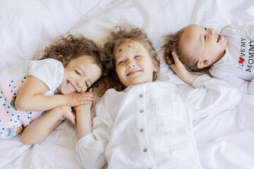three small children lie on white bed and have fun, little boy and two girls smile happy