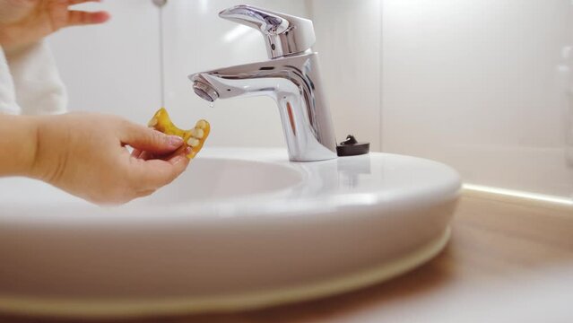 close-up female hand in toilet room, washing wash plastic dentures under running water in white ceramic sink, concept of daily routine hygiene, cleanliness, disinfection protect against bacteria