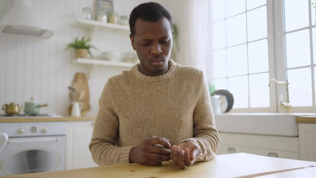 Sad African American man pours vitamin pills from bottle on hand to take in morning. Black guy sits at wooden kitchen table in apartment
