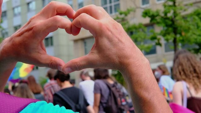 FRANKFURT - July 17, 2021: close-up of male hands in heart form against participants of international LGBT movement, Gay pride parade in city with rainbow flags, mass march of lesbian, gay people