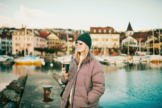 Outdoor portrait of happy young woman drinking takeaway coffee next to lake, image taken in Lausanne, Switzerland
