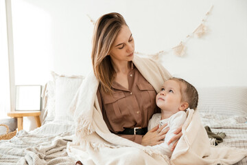 Cute baby girl and her mom enjoy the morning at home in the bedroom. Mom and daughter are sitting on the bed covered with a blanket, covered with a blanket