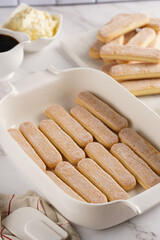 Step by step process of preparing italian dessert tiramisu out of ladyfinger biscuits, mascarpone cheese, espresso on white marble background