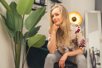 Fototapeta premium Relaxed confident beautiful blonde woman in her 20s or 30s sitting casually and looking at camera in her modern apartment. High quality photo