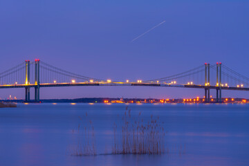 Delaware Memorial Bridge with lights on at dawn colored sky and smooth blue water of Delaware river and reedy grass in foreground.