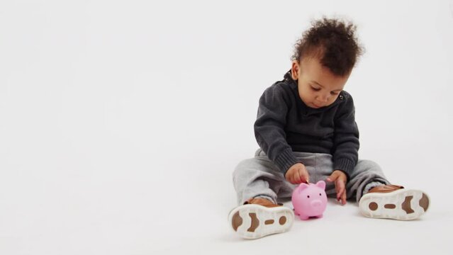 Studio shot of a young baby boy using pink piggy bank by putting coins there over white background.. Savings concept. High quality 4k footage