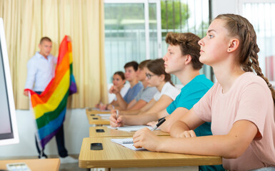 Fototapeta premium Pupils sitting in class and listening carefully to male teacher. He holding LGBT pride flag in hands and talking about sex minorities.