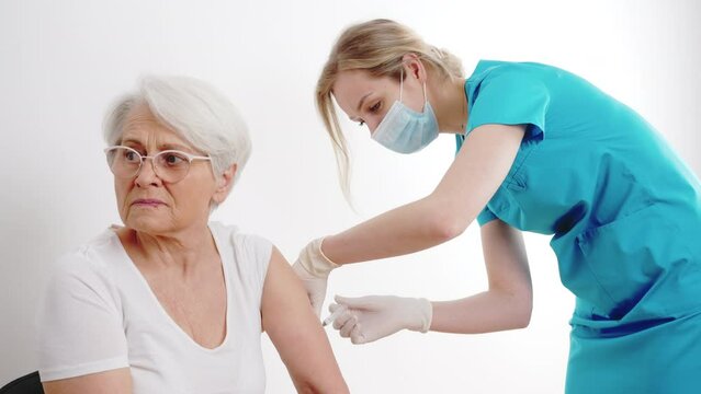 Scared elderly gray-haired grandmother in a white t-shirt is getting vaccinated by healthcare professional worker in a blue outfit. High quality 4k footage