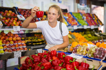 Fifteen-year-old girl who came to the supermarket for shopping, chooses bell pepper, standing near the counter