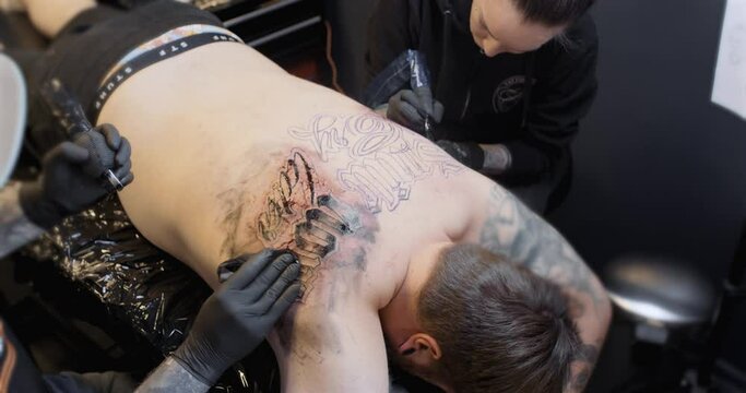 Two tattoo artists working simultaniously on a male back. The closest artist wipes off ink.
4K static shot.