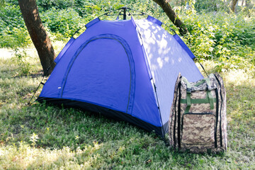 Defocus blue open tourist tent standing on green nature background. Army backpack. Tourism concept. Summer vacation in forest, camping. Blurred. Out of focus