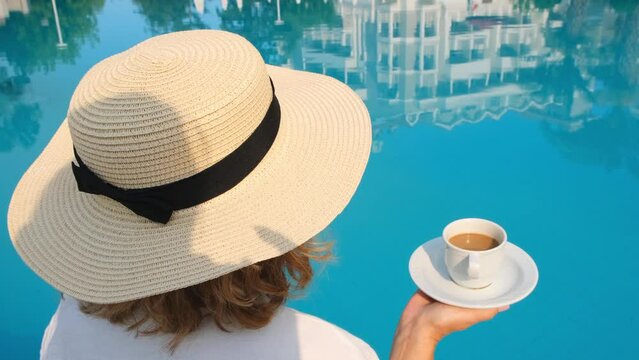 female 50 years old having breakfast by the pool in a straw hat wearing a white dress. woman sitting by the pool with a cup of coffee. good morning and day planning
