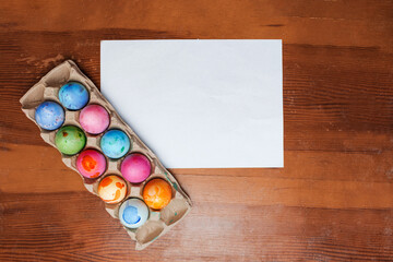 multicolored Easter eggs in a container for eggs are on a wooden old table and next to it lies a white sheet of space for your text. Mockup. view and Selective focus.