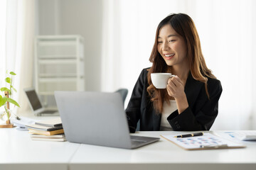 Happy charming asian businesswoman holding a cup of coffee while working on laptop in morning