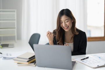 Happy young asian business woman looking at laptop excited by good news online, lucky successful winner man sitting at office desk raising hand in yes gesture celebrating business success win result