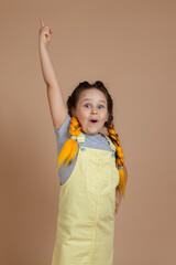 Funny young female with yellow kanekalon pigtails, pointing up with finger looking at camera smiling with opened mouth wearing yellow jumpsuit and gray t-shirt on beige background.