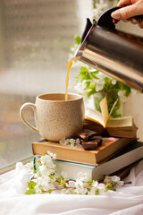 A woman's hand pours coffee into a cup. Composition of books, a cup of coffee and apricot branches on the windowsill.