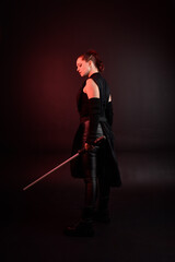 Full length portrait of pretty redhead female model wearing black futuristic scifi leather cloak costume. Standing pose  holding lightsaber on dark studio background with shadow moody lighting.