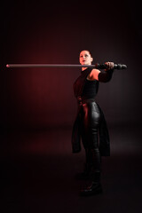 Full length portrait of pretty redhead female model wearing black futuristic scifi leather cloak costume. Standing pose  holding lightsaber on dark studio background with shadow moody lighting.