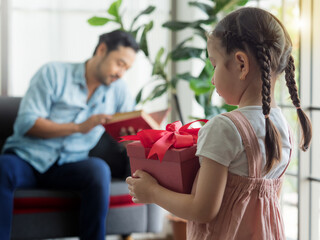 Selective focus, cute daughter holding red box gift with blurry background of father reading book on sofa. Girl want to surprise dad on special day. Father's day or Birthday concept.