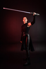 Full length portrait of pretty redhead female model wearing black futuristic scifi leather cloak costume. Standing pose  holding lightsaber on dark studio background with shadow moody lighting.