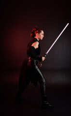 Full length portrait of pretty redhead female model wearing black futuristic scifi leather cloak costume. Standing pose  holding lightsaber on dark studio background with shadow moody lighting.