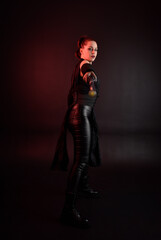 Full length portrait of pretty redhead female model wearing black futuristic scifi leather cloak costume. Standing pose  holding lightsaber on dark studio background with shadow moody lighting.