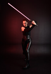 Full length portrait of pretty redhead female model wearing black futuristic scifi leather cloak costume. Standing pose  holding lightsaber on dark studio background with shadow moody lighting.