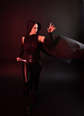 Full length portrait of pretty redhead female model wearing black futuristic scifi leather cloak costume. Standing pose  holding lightsaber on dark studio background with shadow moody lighting.