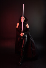 Full length portrait of pretty redhead female model wearing black futuristic scifi leather cloak costume. Standing pose  holding lightsaber on dark studio background with shadow moody lighting.