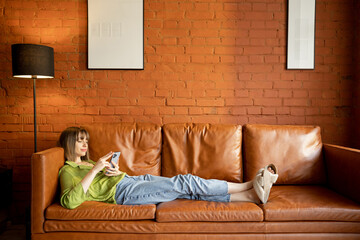 Woman using phone while relaxing on a leather couch at home. Modern Living room with brick wall in loft style. Home coziness and leisure time with gadgets