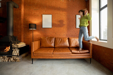 Young carefree woman jumps on a couch, spending time happily at home. Modern living room with fireplace in loft style