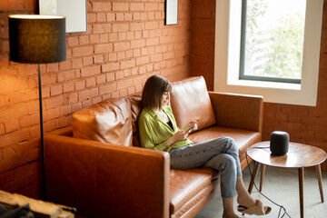 Young woman using phone while sitting relaxed on leather couch with smart speaker in front. Concept...