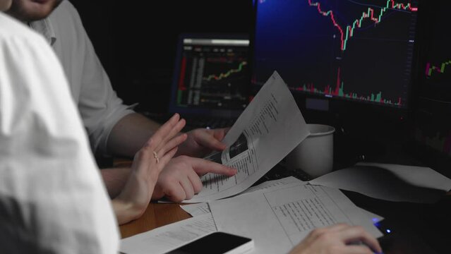 Close-up of the hands of a man and a woman sitting at a computer and looking at an online stock market chart showing bitcoin currencies. Two people compare data from paper and computer. In real time