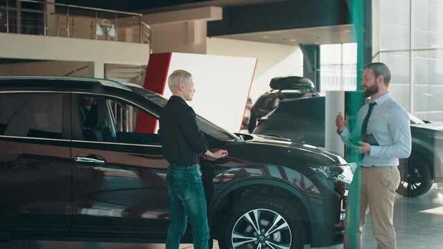 Serious male car seller walking around car salon with blond short hair business woman who wants to buy a car and talking about specifications of cars. Concept of people and transport